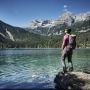 Turystka z plecakiem stoi nad górskim jeziorem w Trentino, w tle widać lasy i skaliste szczyty Dolomitów.