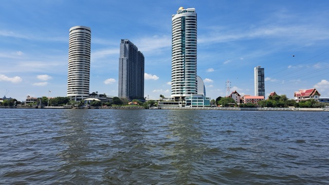 Panorama Bangkoku widziana z rzeki Chao Phraya, z nowoczesnymi wieżowcami i tradycyjnymi świątyniami w tle pod błękitnym niebem.