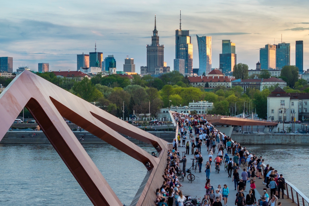 Tłum spacerowiczów i rowerzystów na kładce pieszo-rowerowej nad Wisłą w Warszawie, z panoramą centrum w tle.