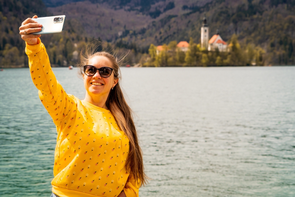 Alt: Kobieta robi selfie nad jeziorem z widokiem na wodę i góry podczas długiego weekendu w czerwcu.