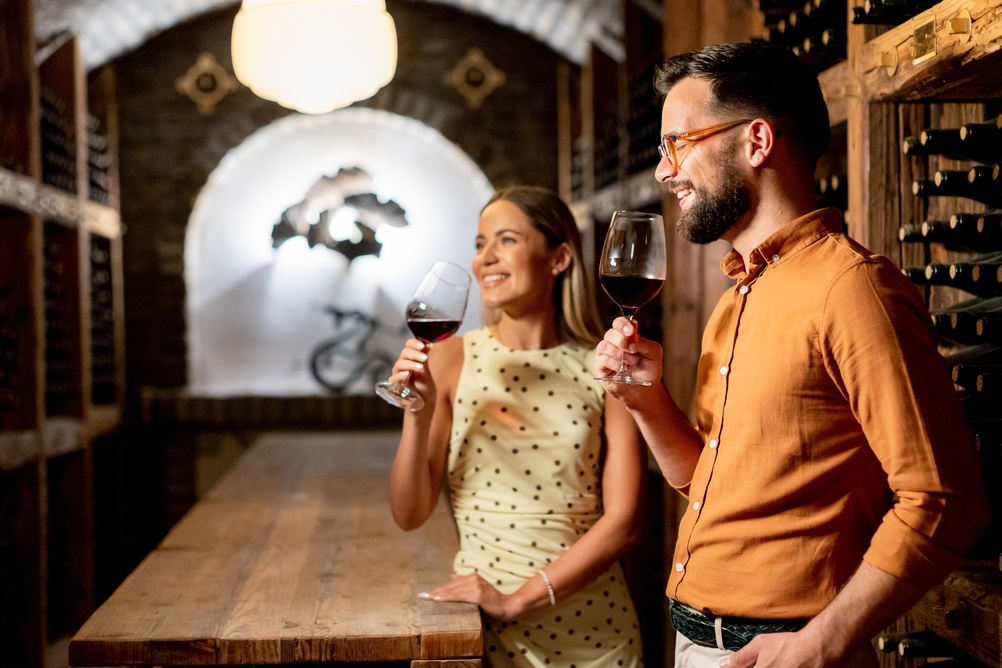 Kobieta i mężczyzna degustują czerwone wino w piwnicy z drewnianymi regałami pełnymi butelek.