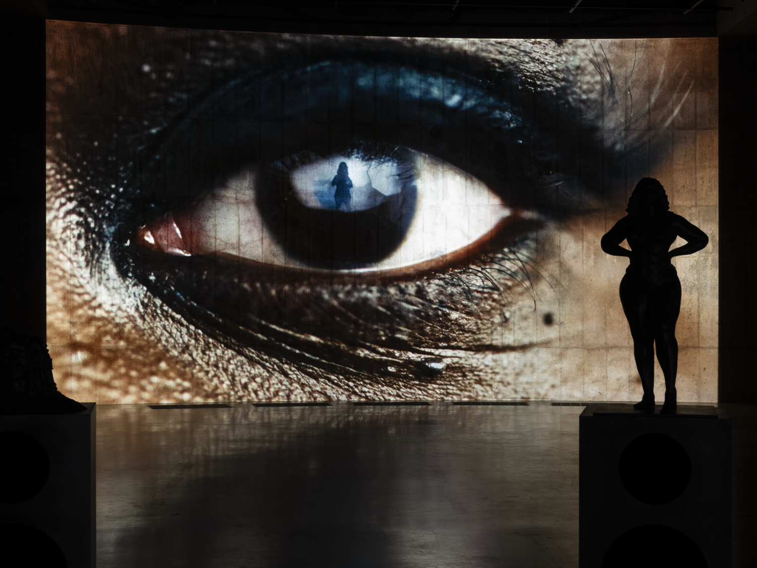 Wielkoformatowa projekcja oka z sylwetką osoby w środku kadru oraz czarna figura stojąca na podeście w ciemnej sali instalacji „Ghost Rider” Christelle Oyiri w Zachęcie.