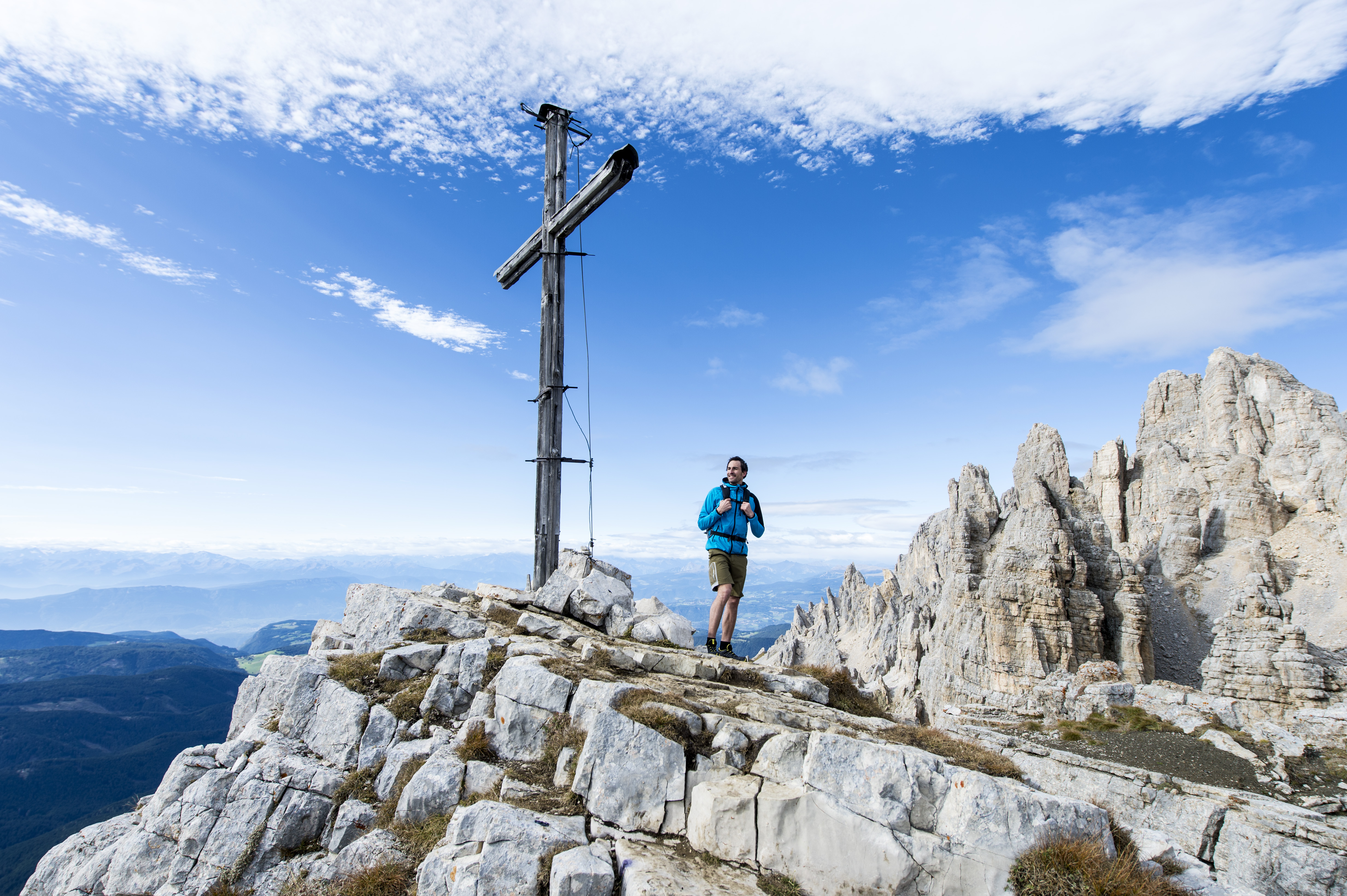 Turysta stojący obok drewnianego krzyża na skalistym szczycie, w tle jasne turnie Dolomitów i rozległa panorama gór.