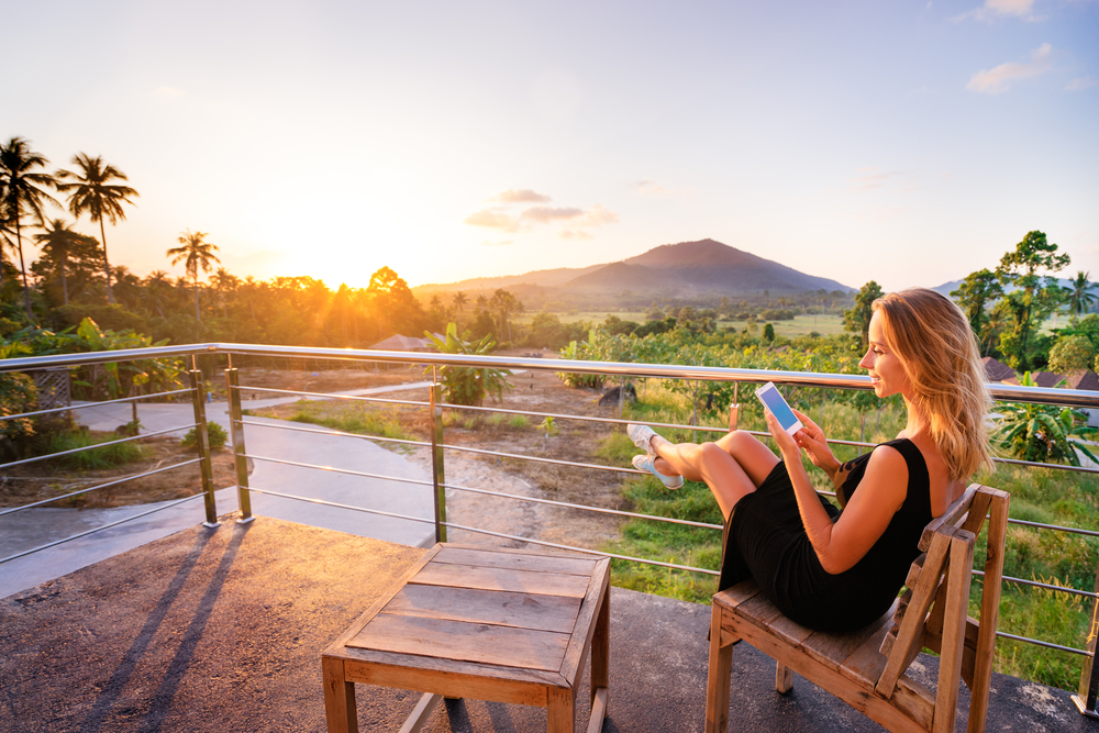 Kobieta na zagranicznych wakacjach siedząca na balkonie z widokiem na palmy i korzystająca z telefonu