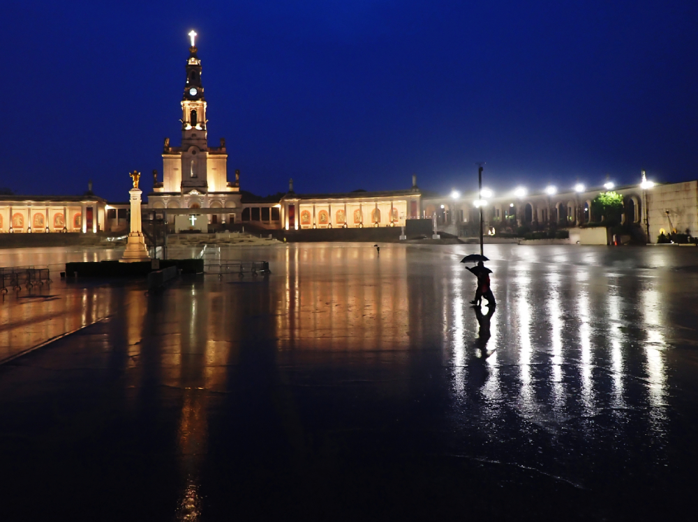 Plac sanktuarium w Fatimie nocą, z oświetloną bazyliką i postacią z parasolem odbijającą się w mokrej nawierzchni.