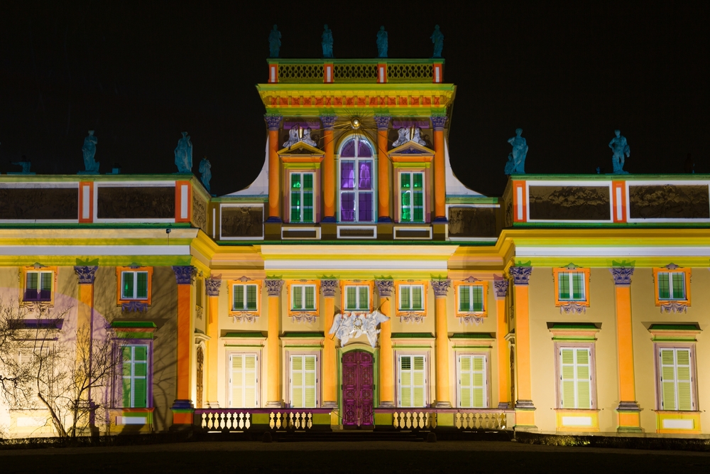 Fasada Pałacu w Wilanowie nocą, oświetlona kolorowymi iluminacjami podkreślającymi kolumny, okna i zdobienia budynku.