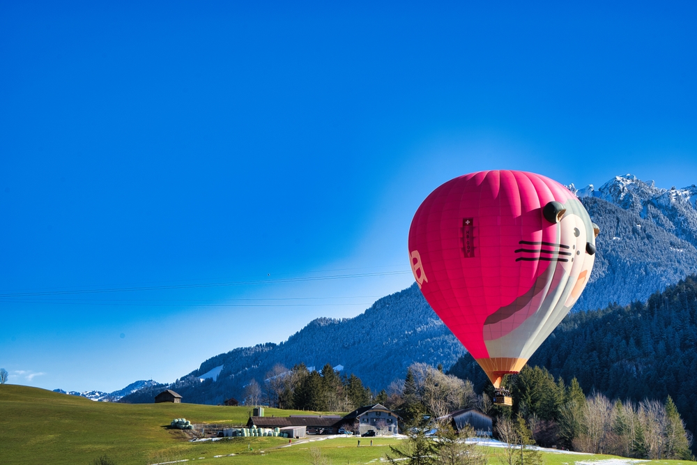 Różowy balon w kształcie głowy kota lecący nisko nad górską doliną, z ośnieżonymi szczytami w tle.