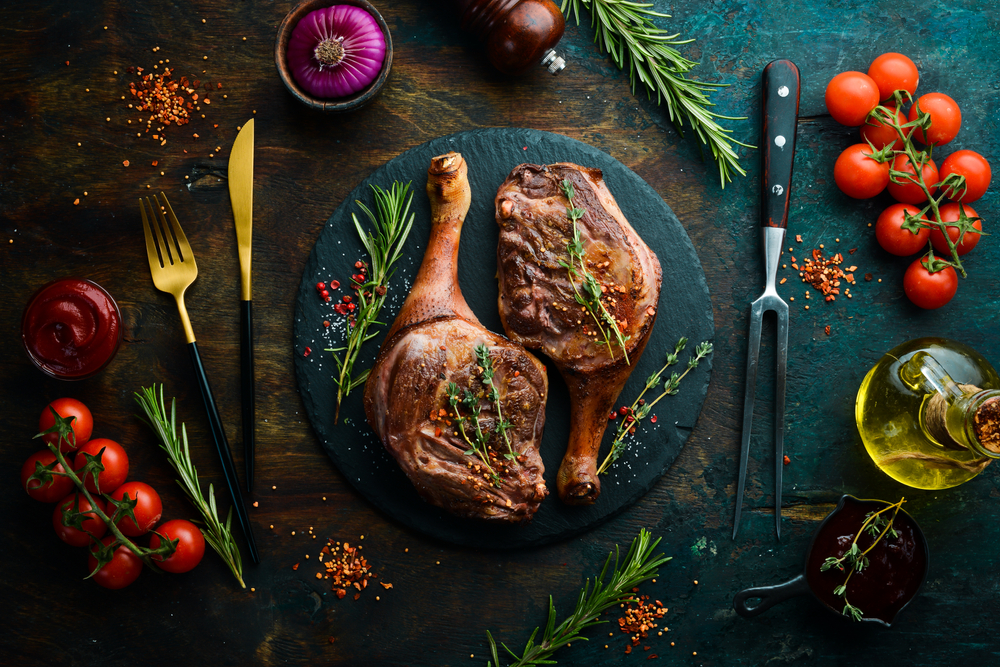 Dwa udka z gęsi z rozmarynem, pomidorami i oliwą na ciemnym tle z dodatkami ziół i przypraw