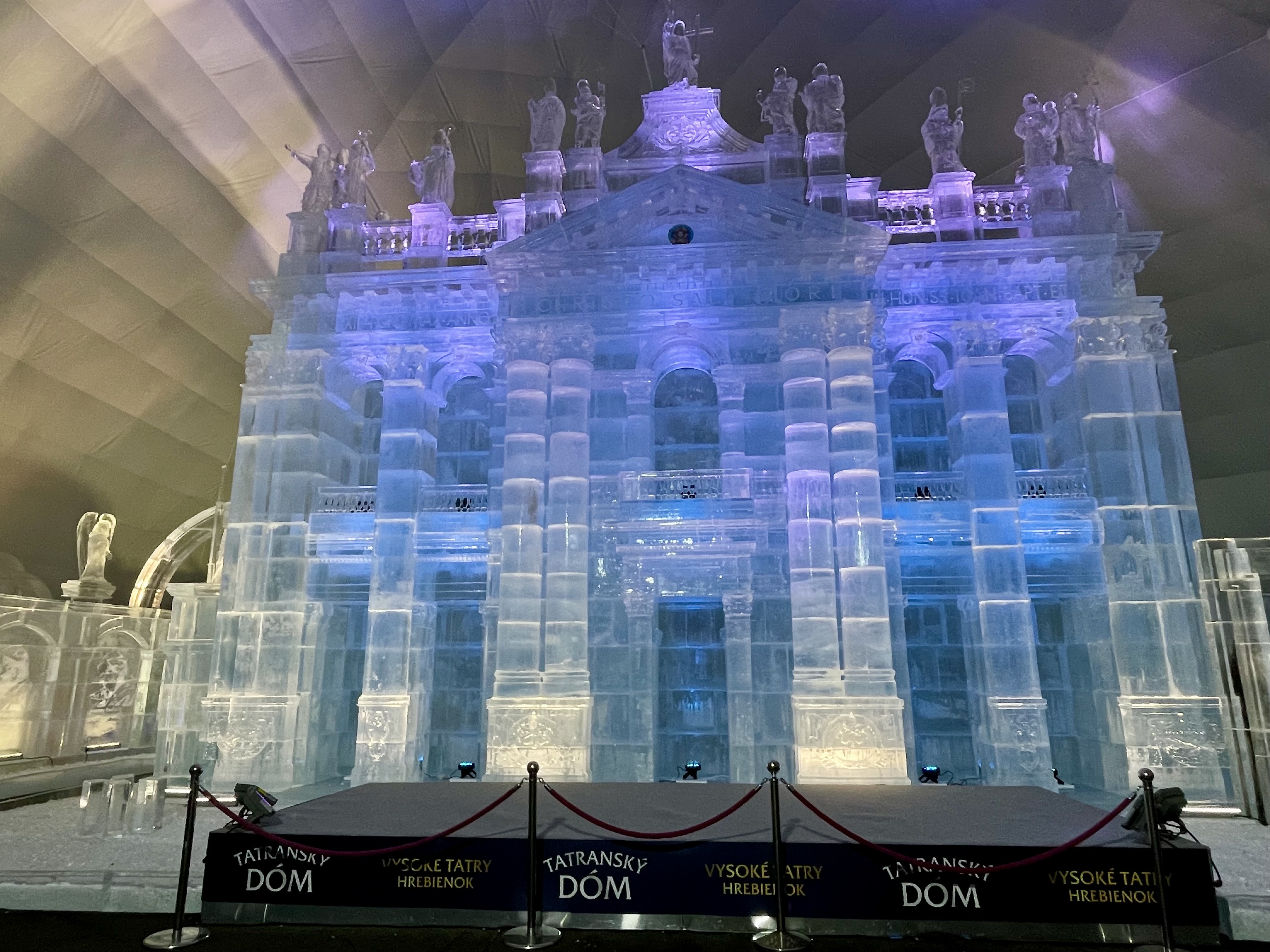 Lodowa fasada świątyni na Hrebienku z wysokimi kolumnami i rzeźbami aniołów, podświetlona na niebiesko i fioletowo.