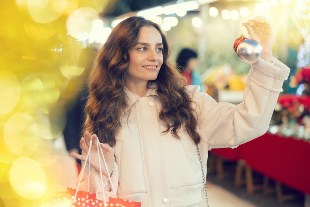 Kobieta oglądająca świąteczne bombki na jarmarku, w tle oświetlone stoiska z ozdobami i dekoracjami.