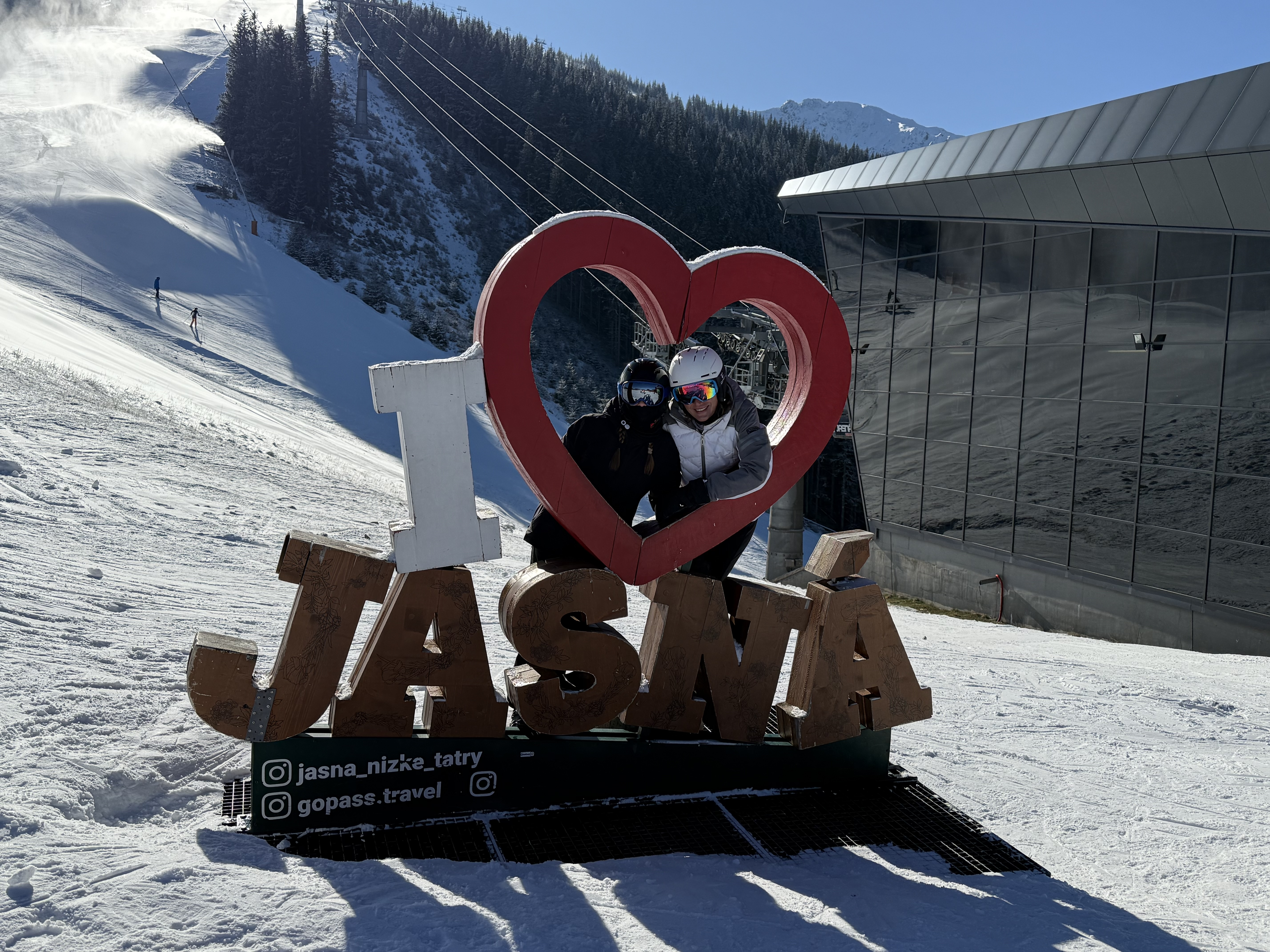 Dwoje narciarzy w kaskach i goglach pozuje w dekoracji „I love Jasná” przy stoku narciarskim i nowoczesnej stacji kolei w Jasnej w Niskich Tatrach.