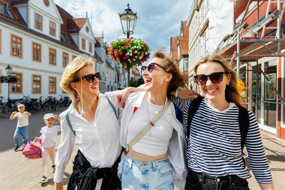 Trzy uśmiechnięte kobiety w okularach przeciwsłonecznych spacerują objęte ulicą z zabytkowymi kamienicami.