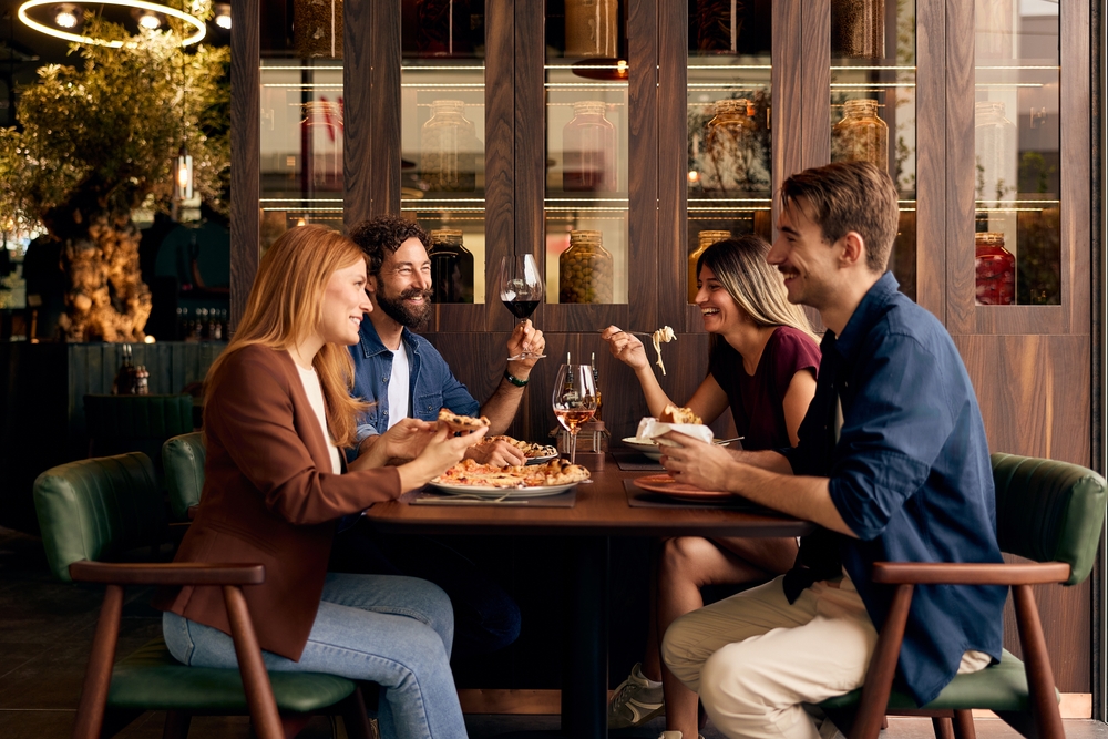 Cztery osoby siedzą przy stole w restauracji, jedzą i rozmawiają w przytulnym wnętrzu z drewnianą zabudową i półkami z przetworami w tle.