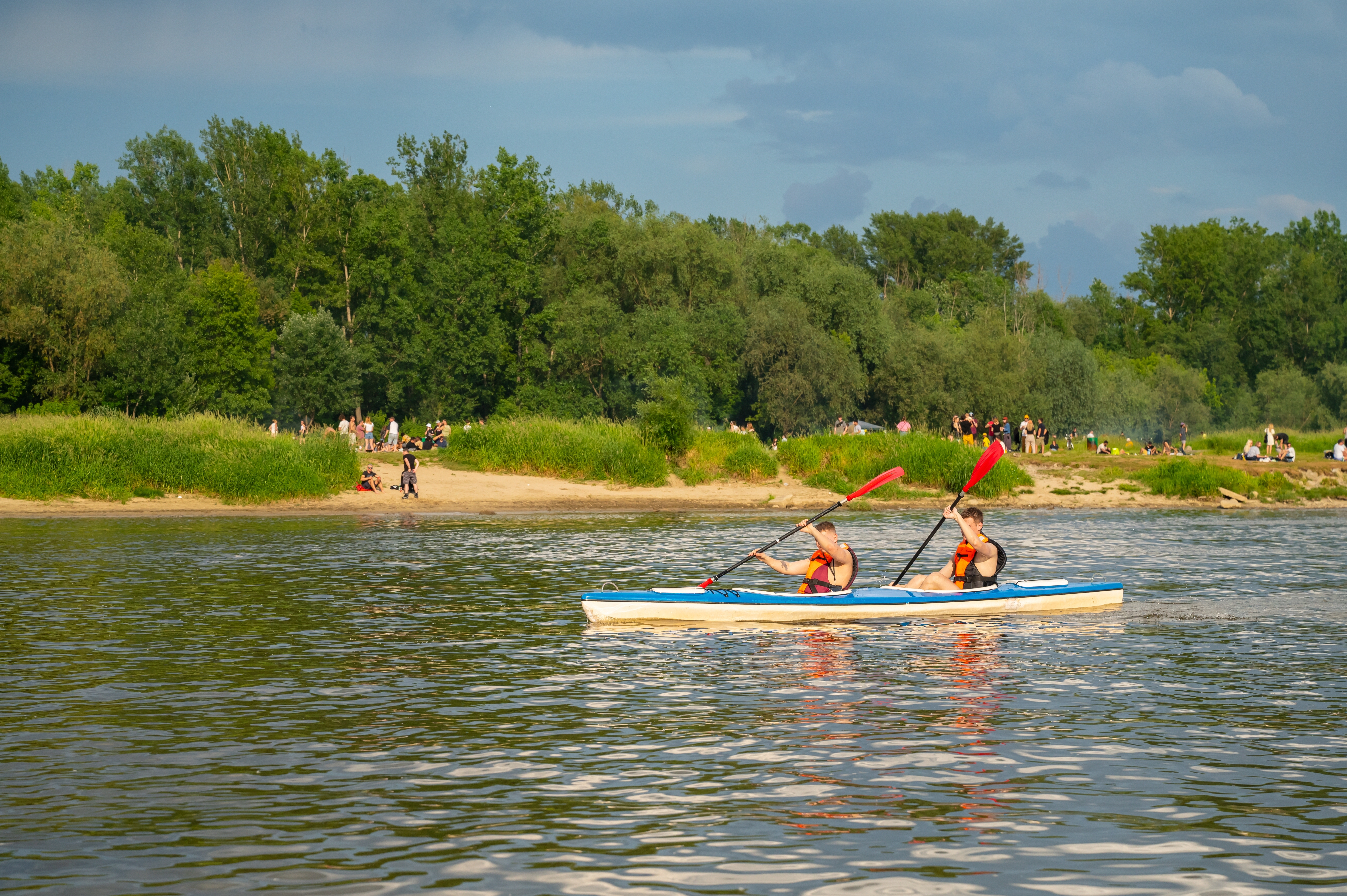 Dwie osoby w kamizelkach ratunkowych płyną dwuosobowym kajakiem po Wiśle, w tle piaszczysta plaża i zielony brzeg.