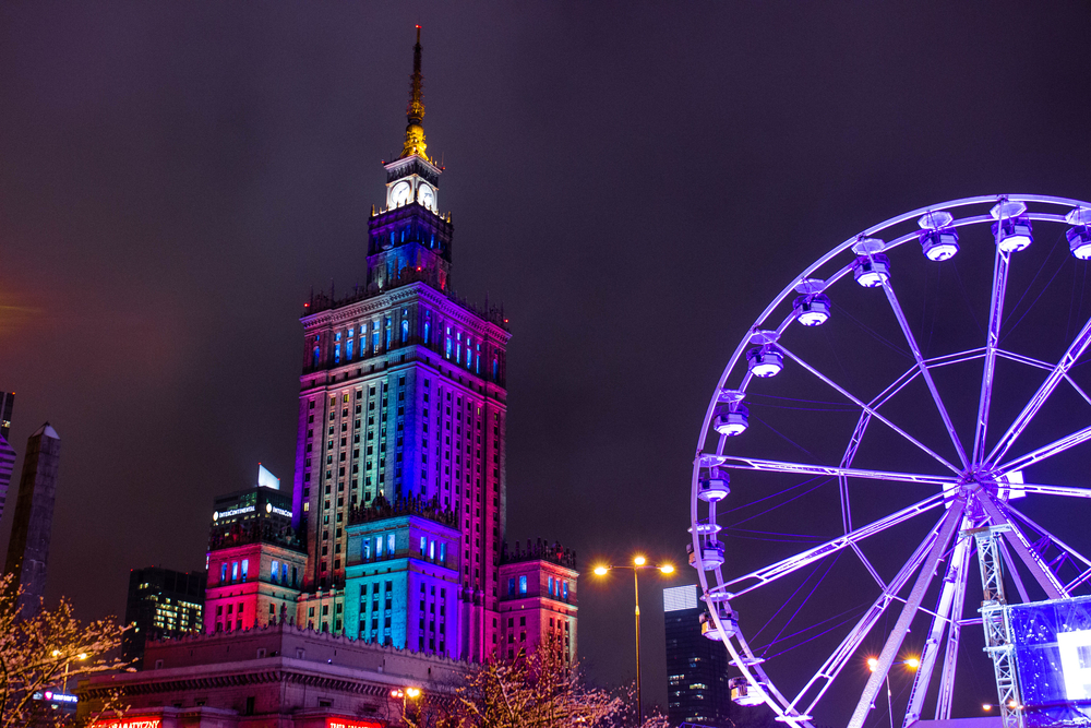 Nocne ujęcie Pałacu Kultury w kolorowych światłach z dużym diabelskim młynem na pierwszym planie.