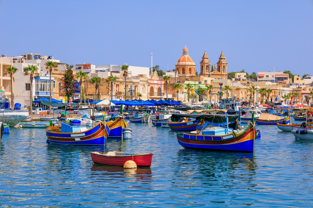 Kolorowe łodzie luzzu w porcie Marsaxlokk na Malcie, w tle zabudowa miasteczka i kościół.
