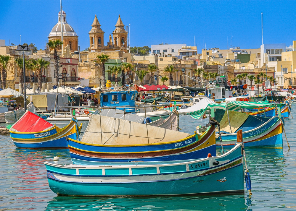 Kolorowe tradycyjne łodzie luzzu w porcie Marsaxlokk na Malcie, zabudowa miasteczka i kościół w tle.