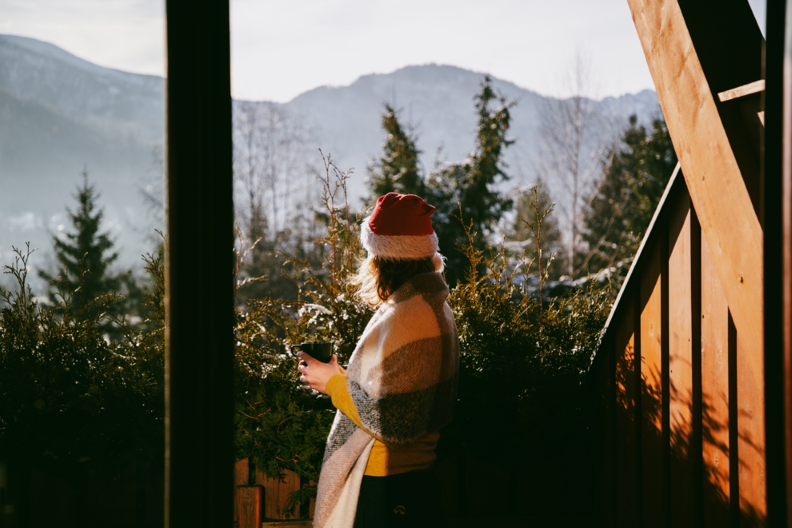 Kobieta w czapce świątecznej i kocu stoi na tarasie górskiego domku z kubkiem w dłoni, w tle zimowe góry