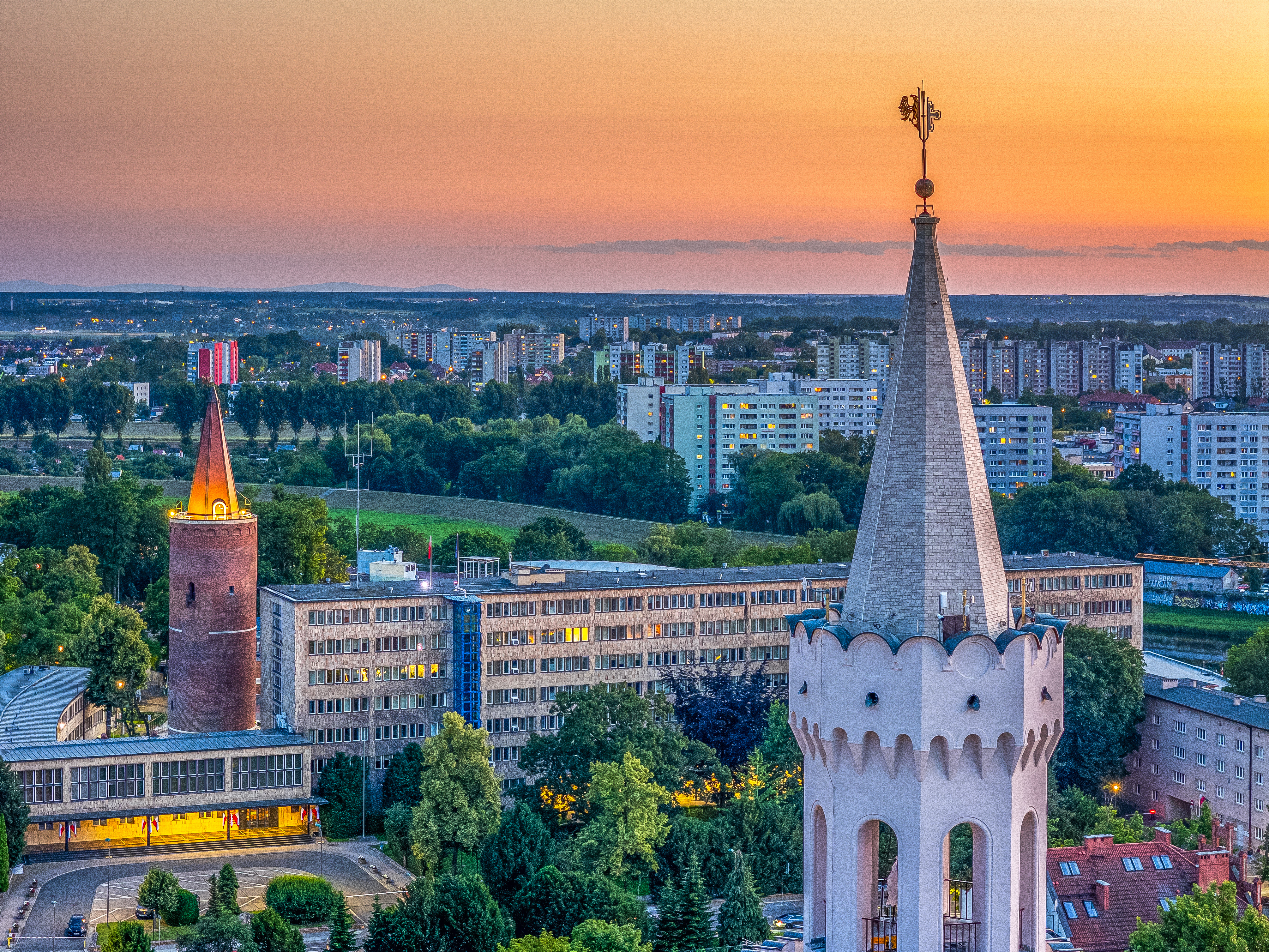 Panorama Opola o zachodzie słońca z miejskimi wieżami, zabudową centrum oraz osiedlami w tle.