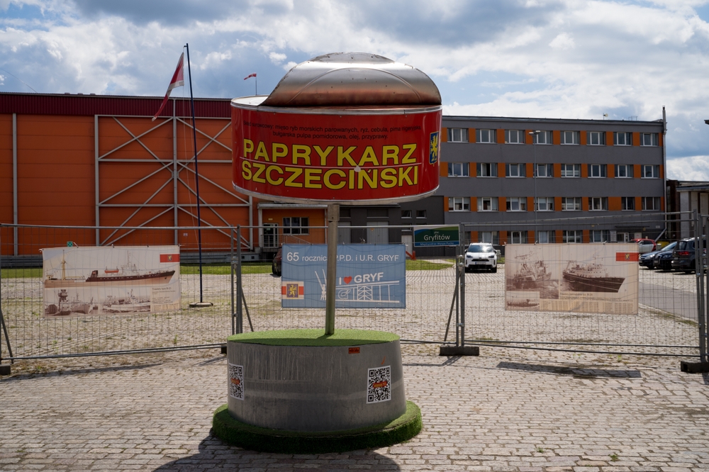 Pomnik Paprykarza Szczecińskiego w formie dużej czerwonej puszki ustawionej na okrągłym postumencie przed zabudowaniami przemysłowymi.