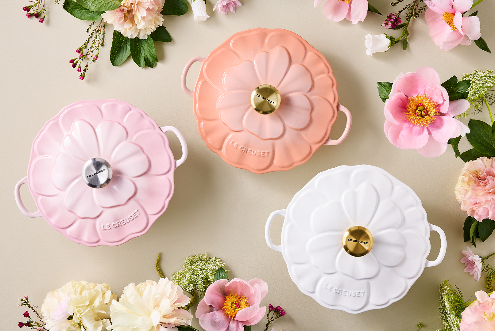 Trzy pastelowe naczynia Le Creuset w kształcie kwiatu, otoczone świeżymi kwiatami na jasnym tle.