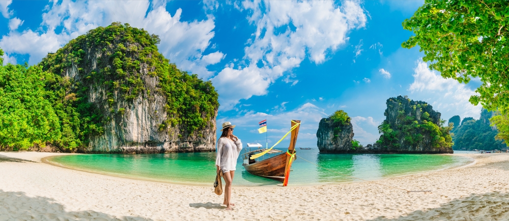 Kobieta na piaszczystej plaży obok tradycyjnej tajskiej łodzi, turkusowa woda i wapienne wyspy Phuket