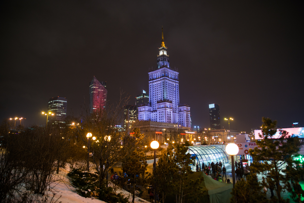 Nocny widok na Pałac Kultury i Nauki w Warszawie oświetlony fioletowym światłem, otoczony drzewami i miejskimi lampami.