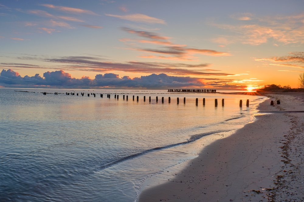 Spokojna plaża nad Bałtykiem o zachodzie słońca, z drewnianymi palami w wodzie i łagodną linią brzegu.