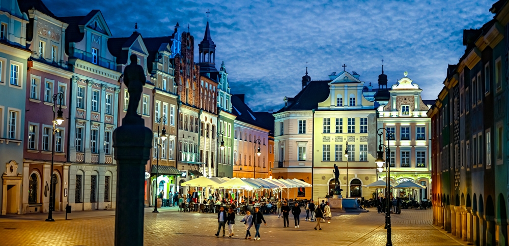 Stary Rynek w Poznaniu nocą z podświetlonymi kamienicami i spacerującymi ludźmi.