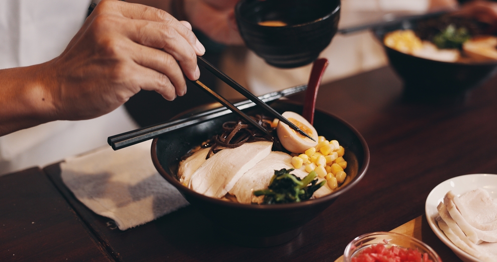 Osoba je ramen pałeczkami z czarnej miski, w której widać plastry mięsa, jajko, kukurydzę i warzywa.