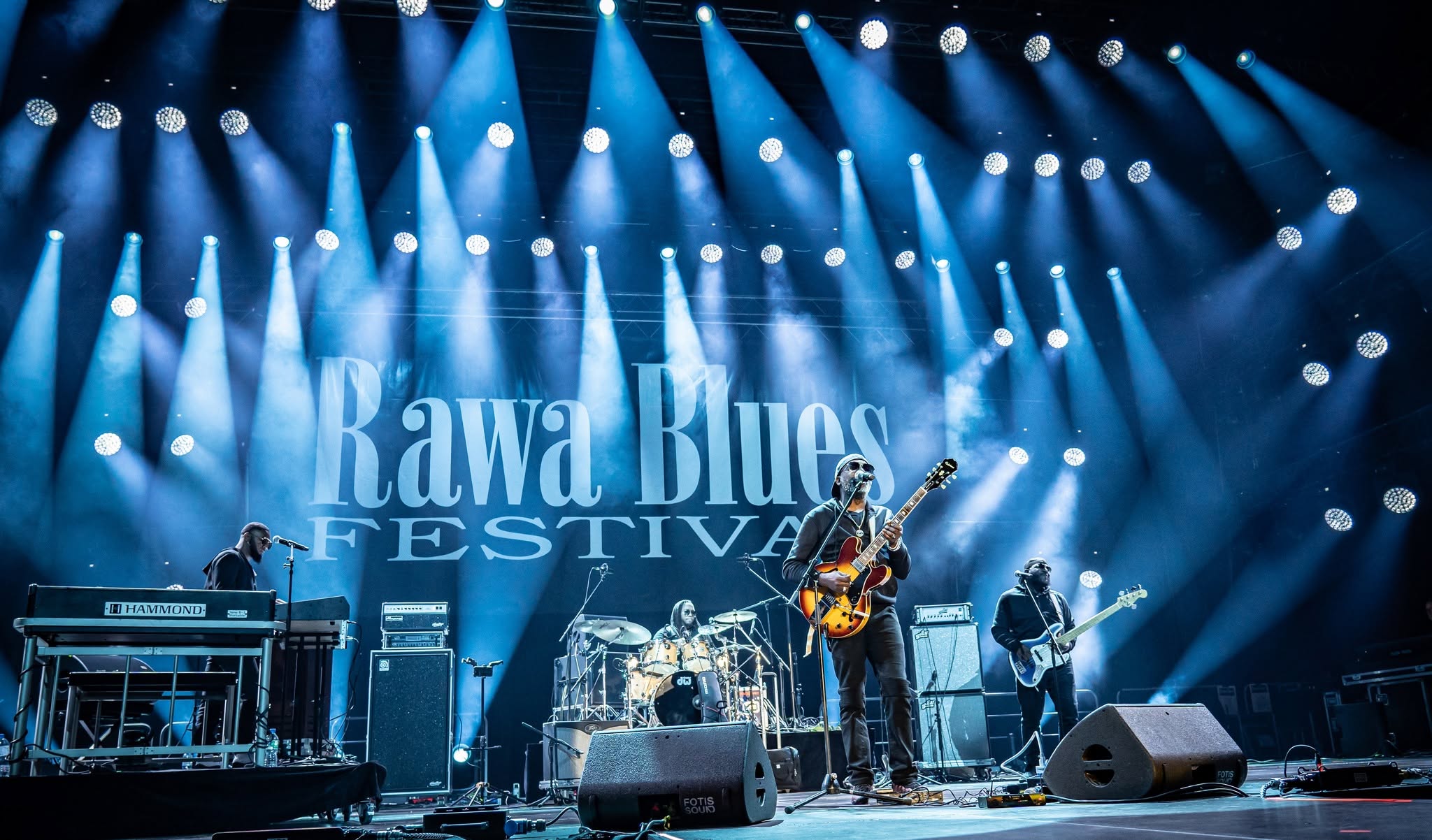 Gitarzysta przy mikrofonie, klawiszowiec, perkusista i basista na scenie Rawa Blues Festival oświetlonej niebieskimi reflektorami.