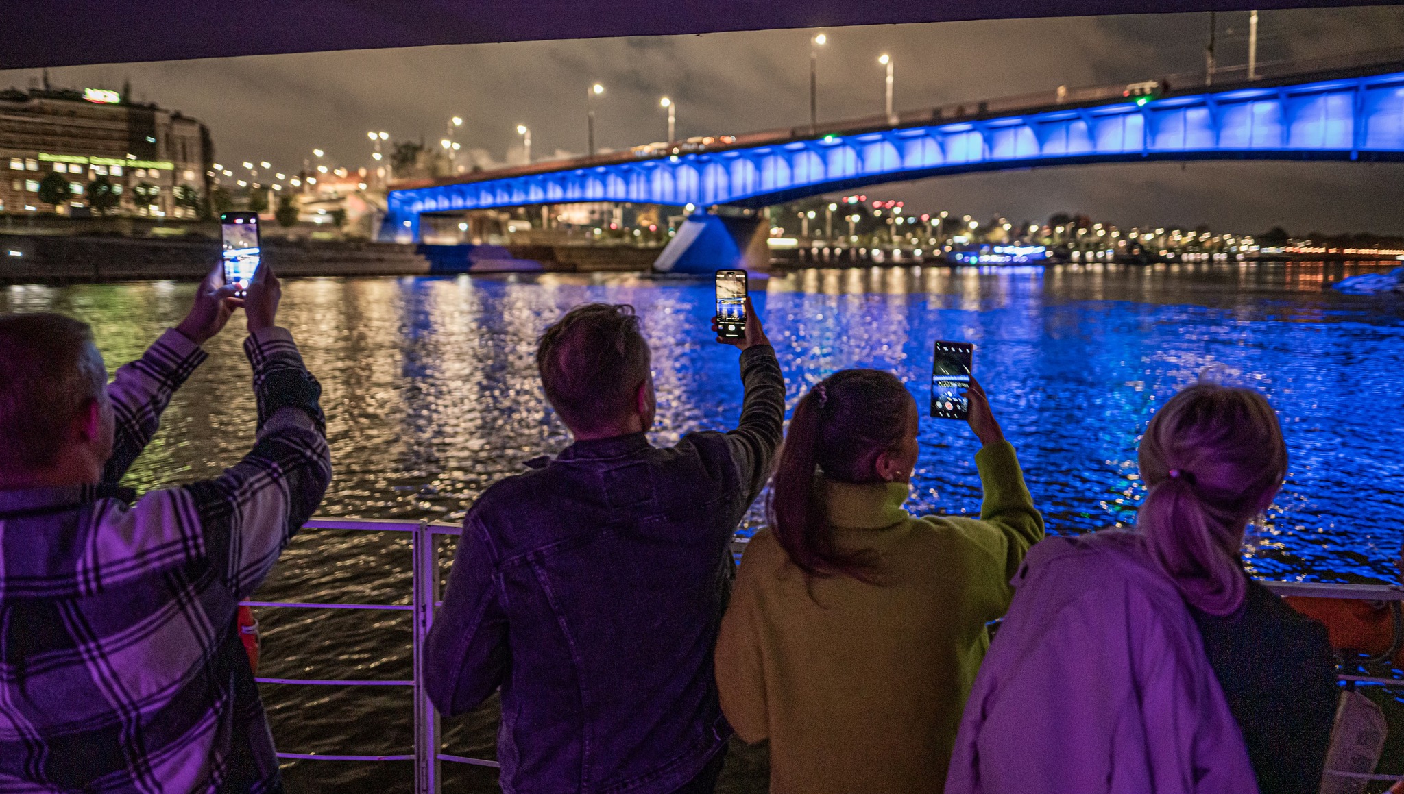 Uczestnicy rejsu fotografują nocną Wisłę i podświetlony most podczas wydarzenia Riverlights w Warszawie.