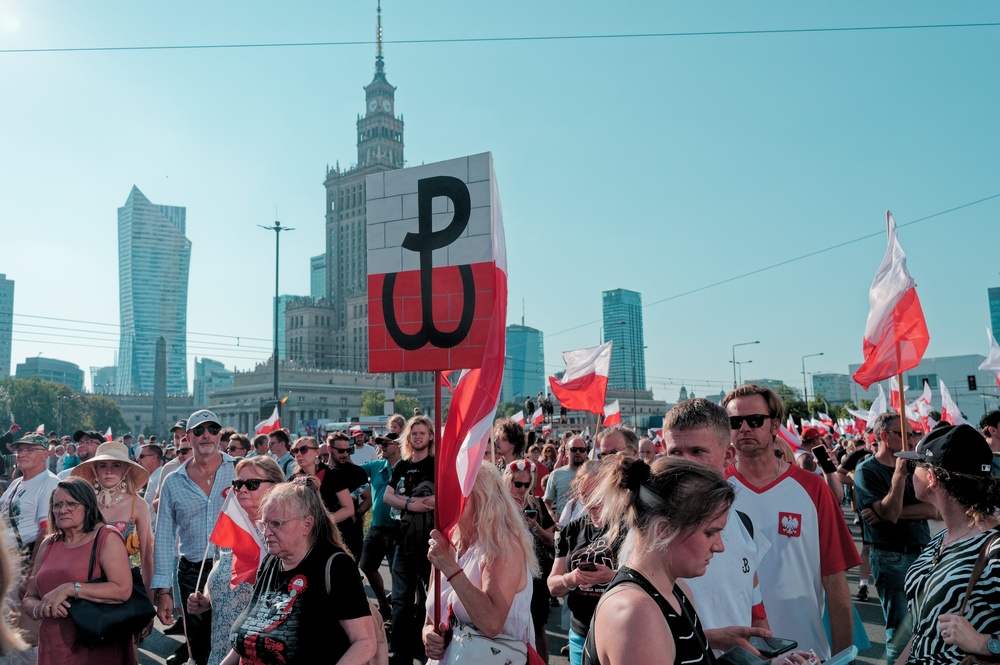 Tłum uczestników obchodów Powstania Warszawskiego z biało-czerwonymi flagami i znakiem Polski Walczącej w centrum Warszawy.