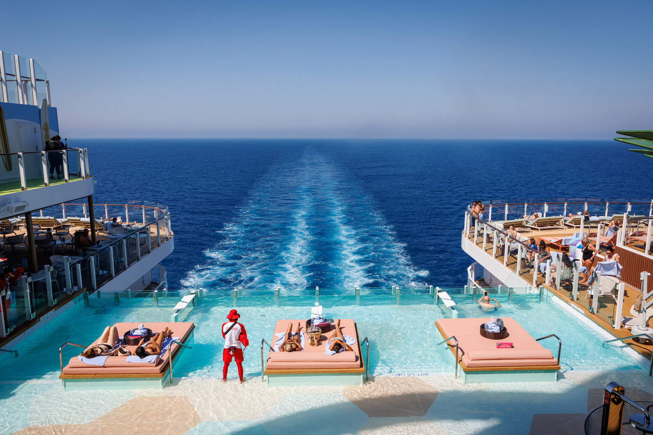 Błękitny basen infinity w strefie The Hideaway na rufie Legend of the Seas, z leżankami ustawionymi w wodzie i widokiem na morze oraz ratownikiem stojącym w wodzie.