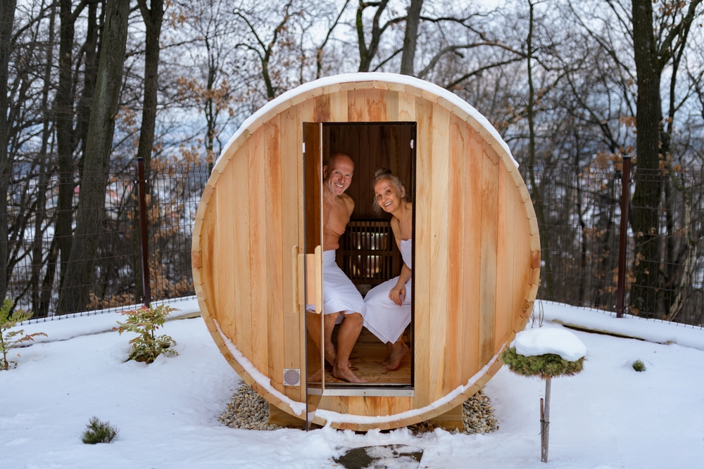 Para siedząca w drewnianej saunie-beczce w zimowej scenerii, śnieg i drzewa wokół.