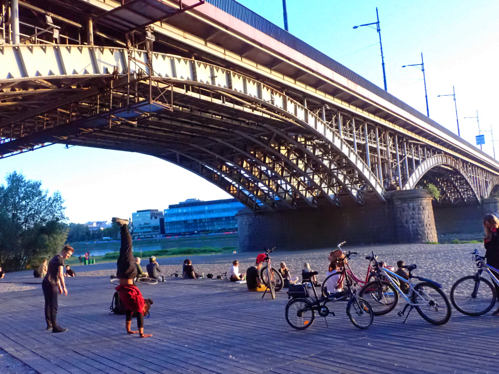Praski brzeg Wisły w Warszawie pod mostem Poniatowskiego z rowerami, plażą i odpoczywającymi ludźmi.