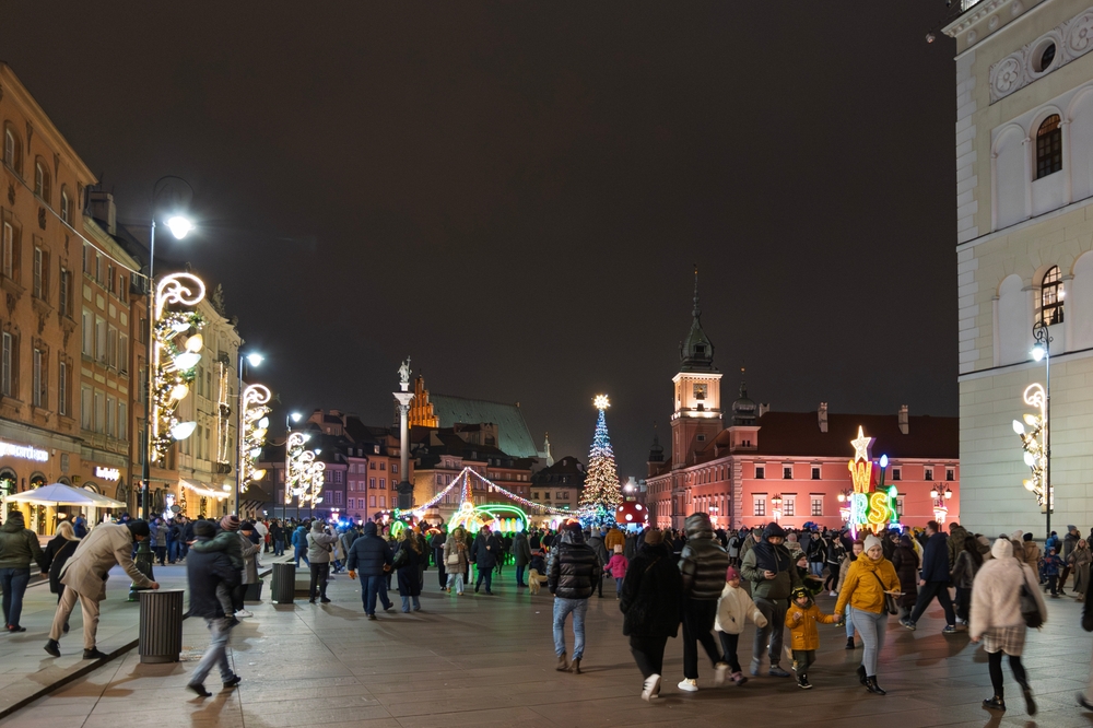Świąteczny Plac Zamkowy w Warszawie nocą: choinka, iluminacje i spacerujący ludzie na starówce zimą.