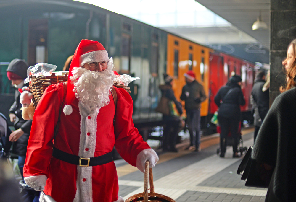 Mikołaj z koszem prezentów na peronie obok świątecznego pociągu w kolorowych wagonach, w tle pasażerowie w zimowych ubraniach.