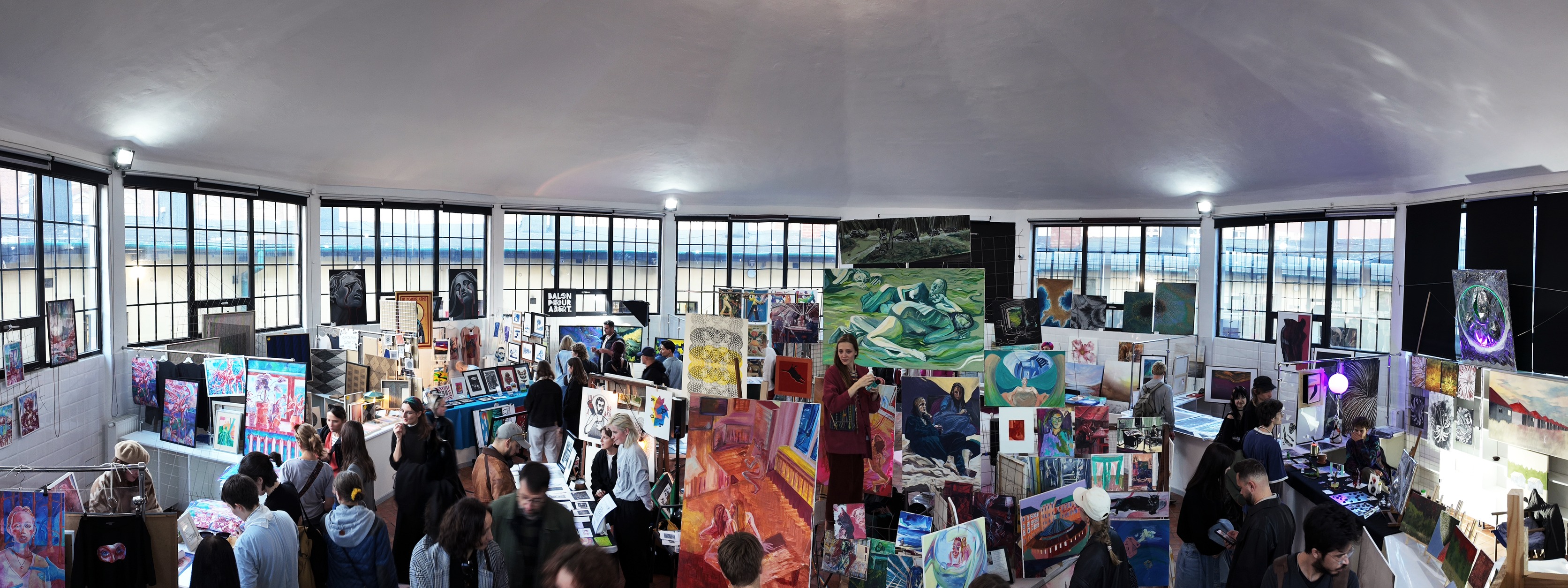 Odwiedzający oglądają obrazy i ilustracje na Targach Młodej Sztuki w jasnej hali wystawowej