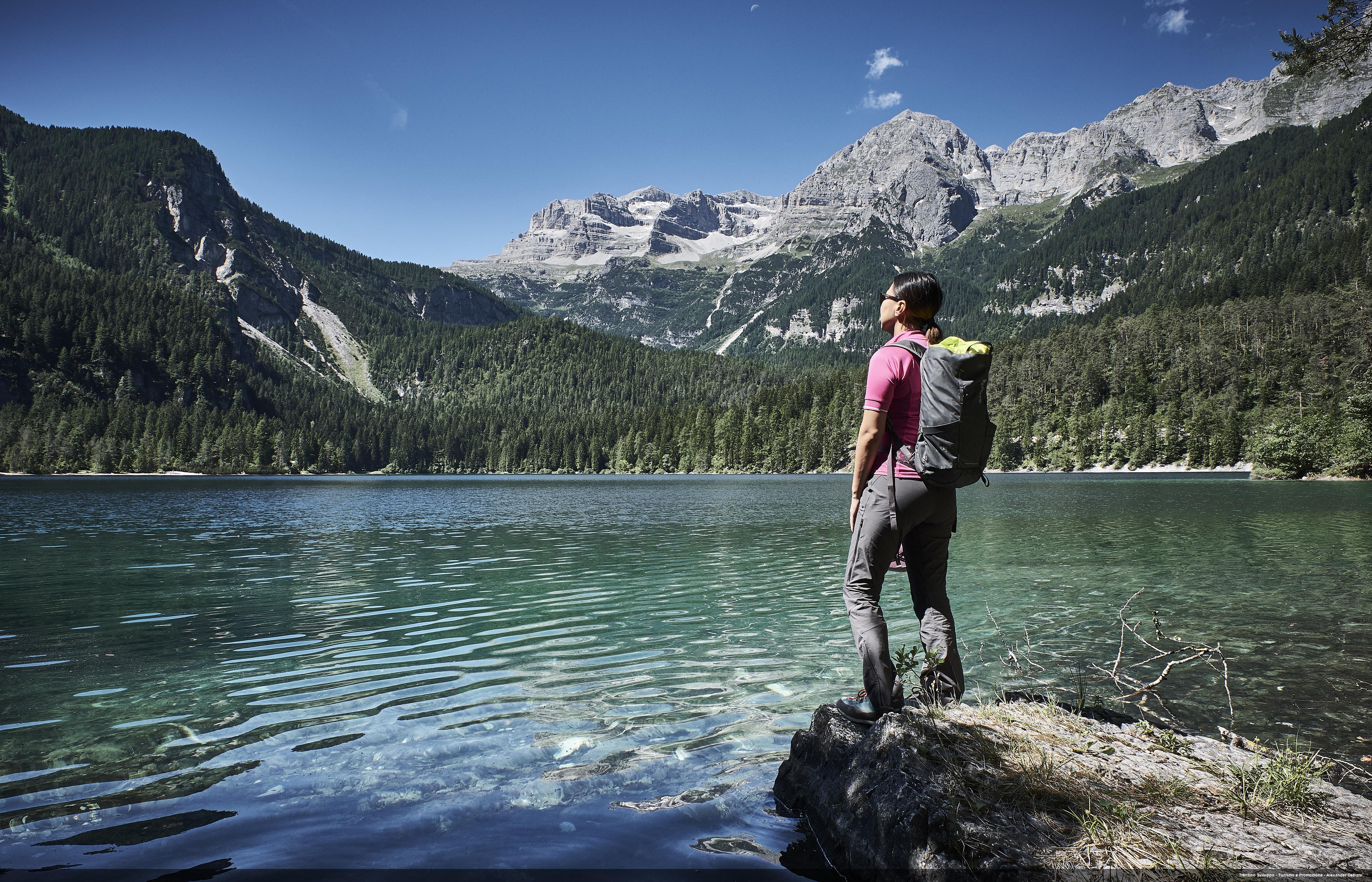 Turystka z plecakiem stoi nad górskim jeziorem w Trentino, w tle widać lasy i skaliste szczyty Dolomitów.