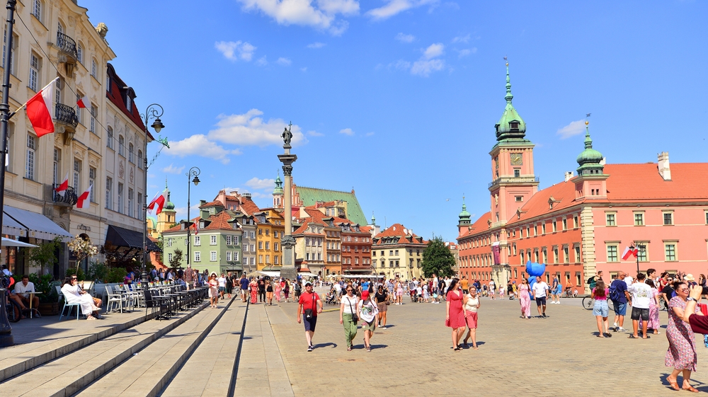 Plac Zamkowy i Stare Miasto w Warszawie w słoneczny dzień, spacerujący mieszkańcy i turyści.