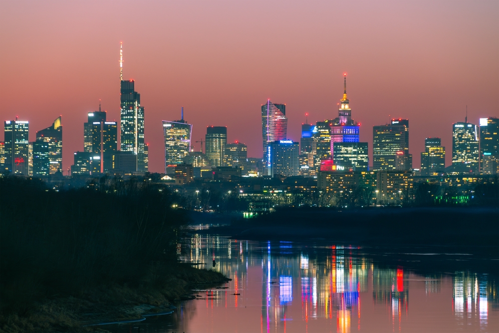 Panorama centrum Warszawy nocą z widokiem na Pałac Kultury i Nauki i wieżowce odbijające się w Wiśle.