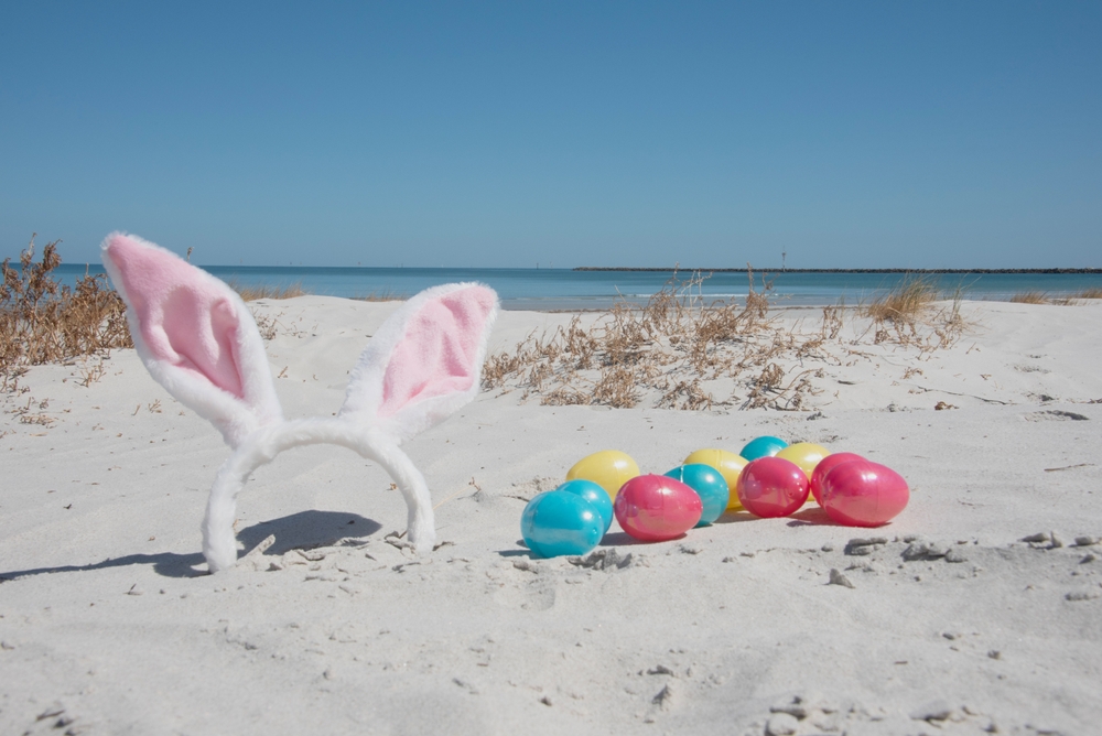 Kolorowe wielkanocne jajka i opaska z uszami królika ustawione na jasnym piasku przy plaży nad morzem.