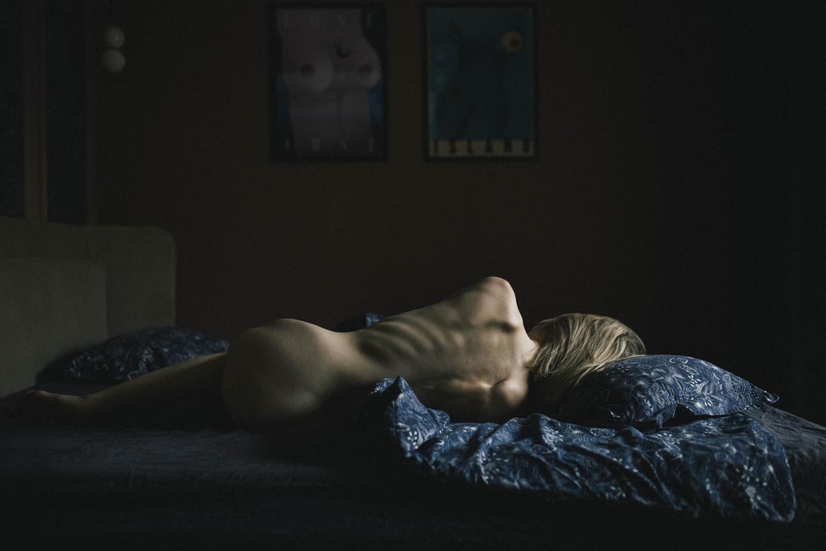 Kobieta leży na łóżku w ciemnym pokoju, odwrócona plecami do obiektywu, oświetlona wąskim, przygaszonym światłem. Fot. Katarzyna Żak